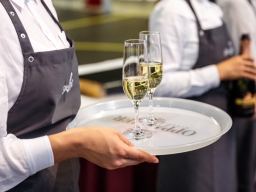 Service Ein Gastromitarbeiter trägt zwei Gläser mit Sekt auf einem Tablett. Im Hintergrund steht ein Mitarbeiter mit einer geöffneten Sektflasche.