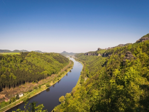 View of the Elbe River downstream toward Lilienstein. The banks of the Elbe are green, the sky blue, and there are no clouds in sight. To the left is the railway line to Dresden, but there are no trains running at the moment.