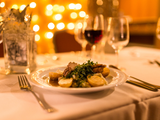 On a set table stands a plate of roast goose In the background, several wine glasses stood on a set table, one of which is filled with red wine. On a plate in the foreground, a piece of roast goose with gravy, vegetables, and potatoes lies.